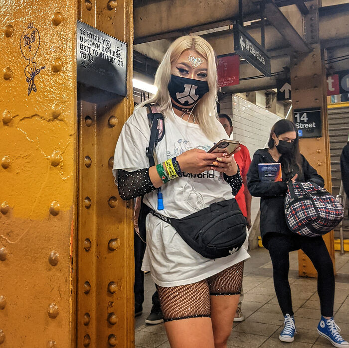 Woman with face mask and glitter makeup using phone, wearing fishnet tights, standing in NYC subway station portrait.