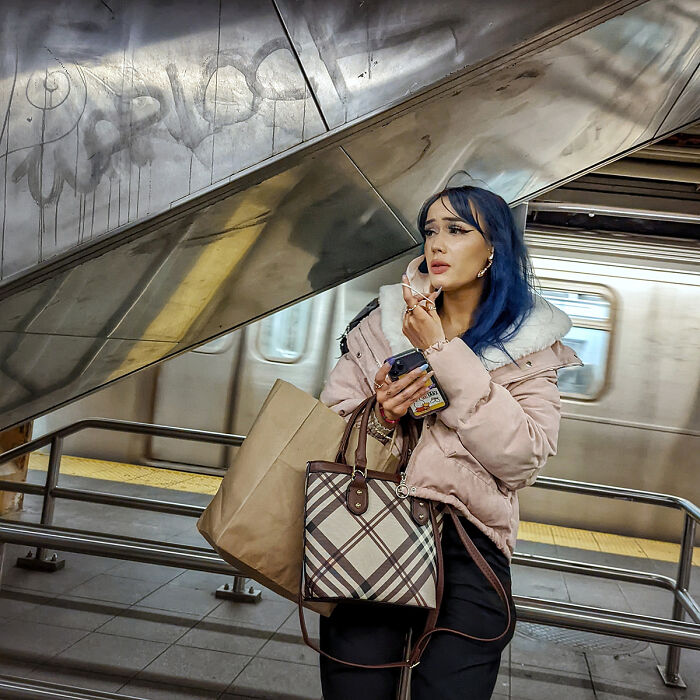 Young woman with blue hair on a phone call, holding bags, waiting near subway platform railing in NYC subway station.