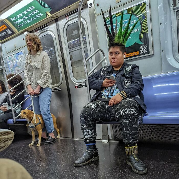Punk passenger with green mohawk hairstyle sitting in NYC subway, holding phone, capturing street portraits underground.