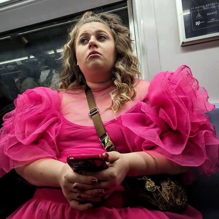 Woman wearing a bright pink ruffled dress holding phone, captured in a captivating street portrait inside NYC’s subway.