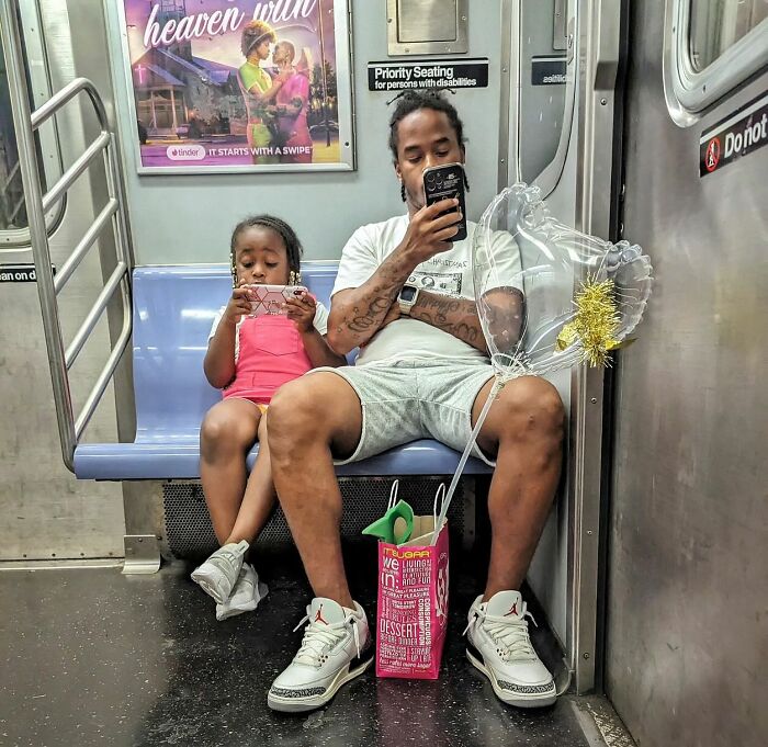 Father and daughter seated in NYC subway, each focused on their phones, with a heart-shaped balloon nearby in street portrait style.