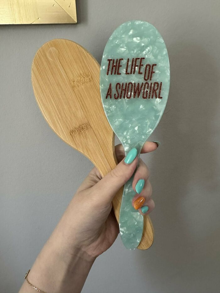 Hand holding a teal hairbrush with The Life of a Showgirl text alongside a wooden hairbrush, Taylor Swift merchandise. Hand holding a teal hairbrush with The Life of a Showgirl text alongside a wooden hairbrush, Taylor Swift merchandise.