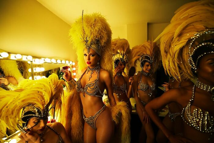Showgirls in elaborate feather costumes preparing backstage with bright dressing room lights and mirrors behind them. Showgirls in elaborate feather costumes preparing backstage with bright dressing room lights and mirrors behind them.