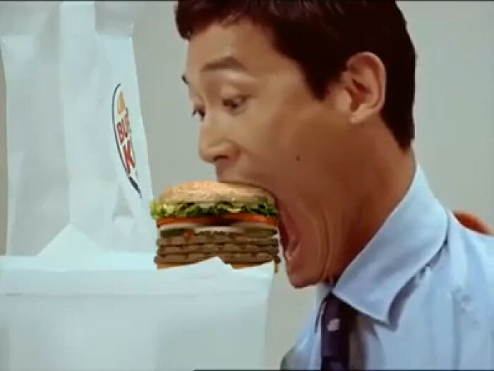 Man with an exaggeratedly large mouth about to eat a massive burger in a creepy commercial scene.