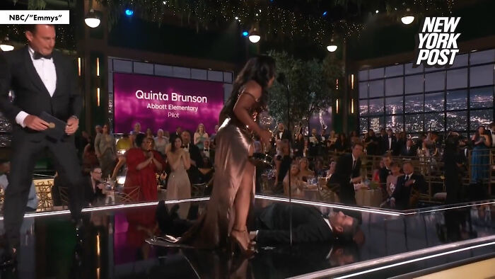 Woman in a shiny dress helping a man lying on the ground at an awards event, capturing a notable oopsie moment.