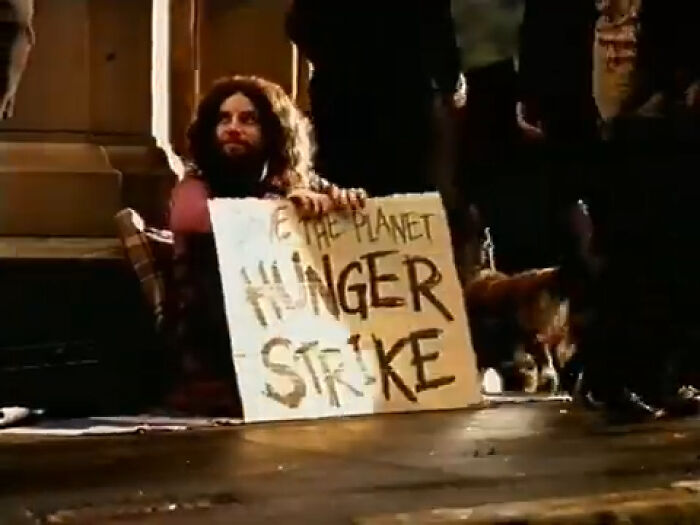 Man with long hair sitting on the ground holding a cardboard sign about hunger strike in a creepy commercial scene at night.