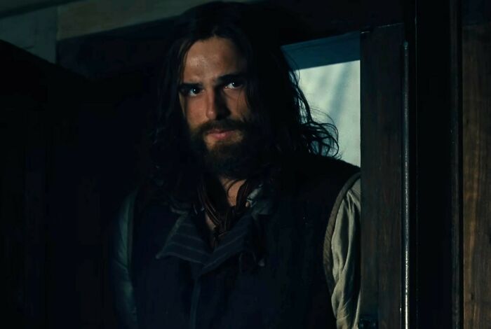 A man with long hair and beard in period costume standing in a dimly lit room from Wuthering Heights trailer. A man with long hair and beard in period costume standing in a dimly lit room from Wuthering Heights trailer.
