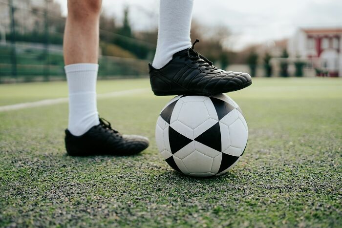 Close-up of a person wearing black cleats with one foot resting on a soccer ball on a school sports field.