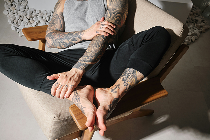 Man with detailed arm and leg tattoos, wearing a gray tank top and black pants, sitting cross-legged on a chair. Man with detailed arm and leg tattoos, wearing a gray tank top and black pants, sitting cross-legged on a chair.