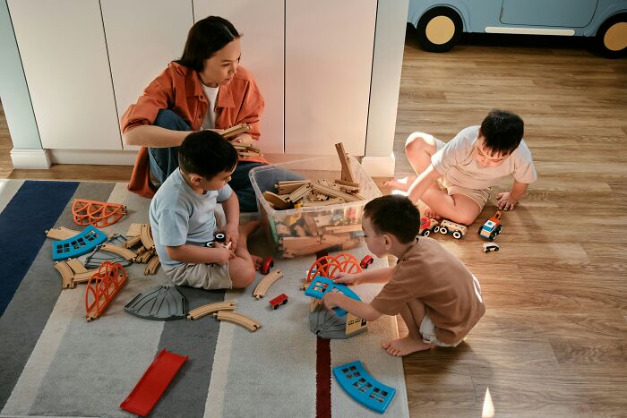 Woman and children playing with wooden train tracks at home, illustrating dark secrets that could ruin lives if revealed.