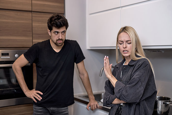 Wife standing upset with arms crossed as husband looks on in kitchen, highlighting struggles with bed cleaning and refusal to use aid. Wife standing upset with arms crossed as husband looks on in kitchen, highlighting struggles with bed cleaning and refusal to use aid.