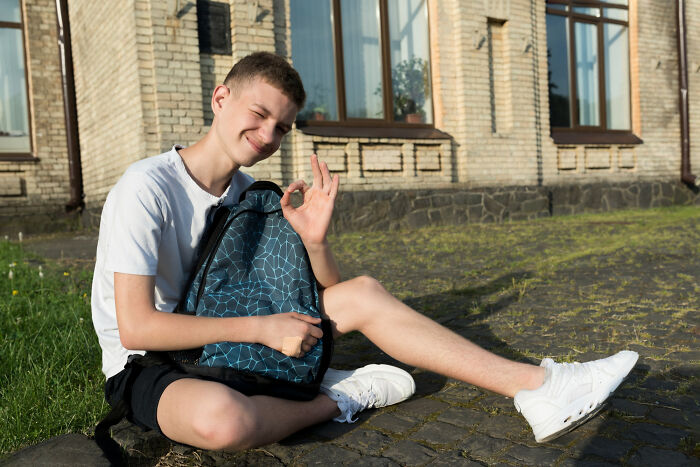 Teenage boy sitting outside a house, holding a backpack and smiling, symbolizing family relationship and home connection.