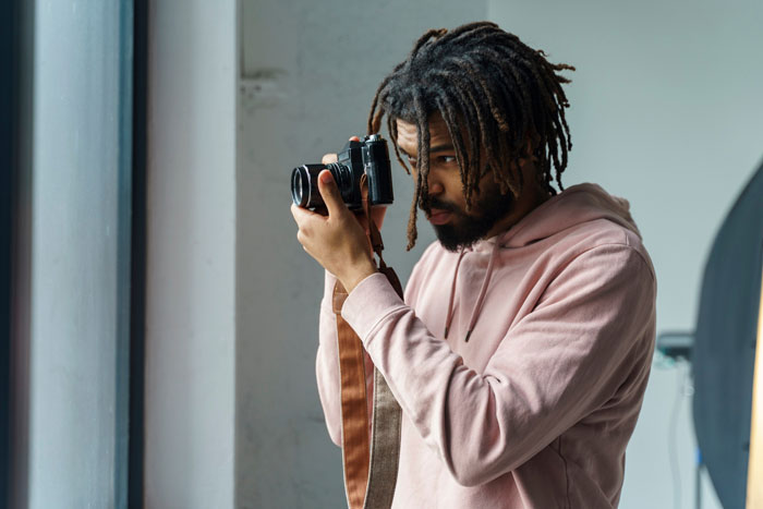 Photographer with dreadlocks and a pink hoodie focusing through the camera lens capturing indoor pictures.
