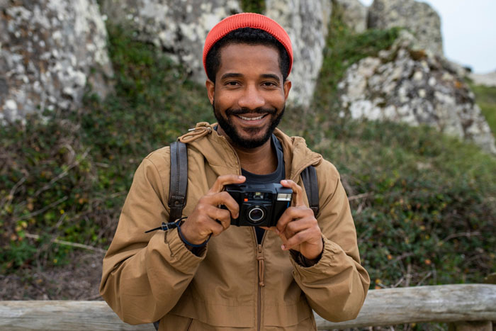 Smiling photographer outdoors wearing a red beanie and tan jacket, holding a camera near rocky and green natural background.