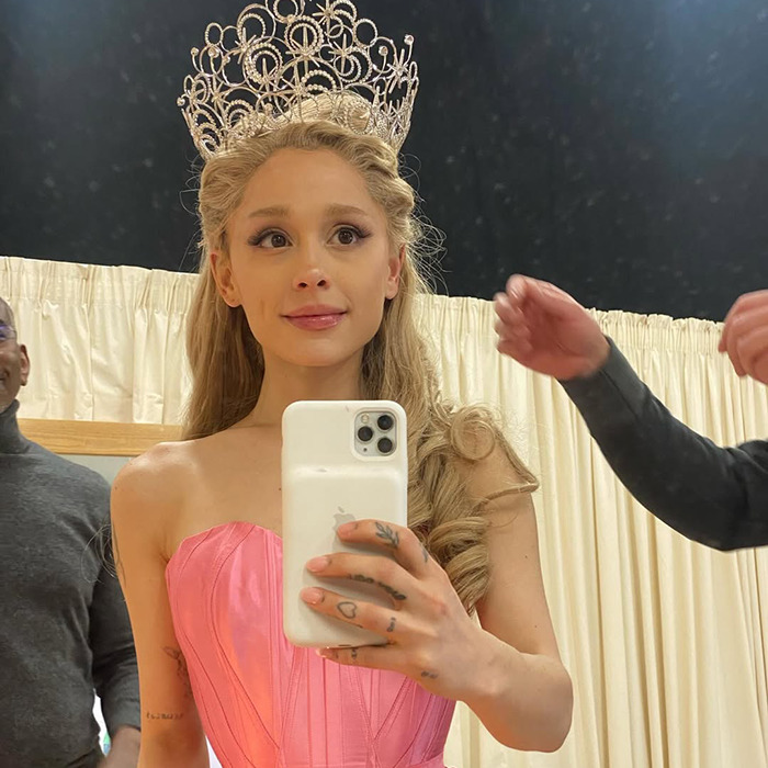 Ariana Grande taking a selfie wearing a pink dress and intricate crown, showing her dramatic new look in a backstage setting. Ariana Grande taking a selfie wearing a pink dress and intricate crown, showing her dramatic new look in a backstage setting.