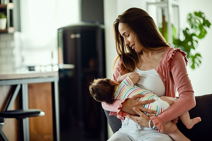 Young mother gently cradling newborn baby in living room, highlighting family dynamics after birth with stepsister present.