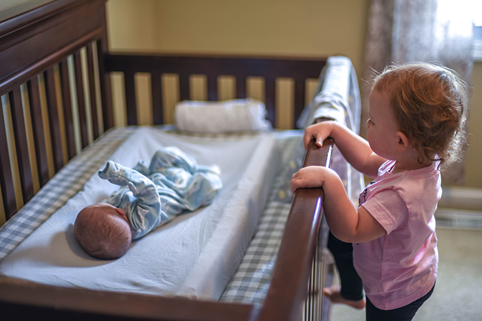 Toddler sister watching baby brother sleeping in crib, revealing dead brother lie emotions in a home nursery setting.