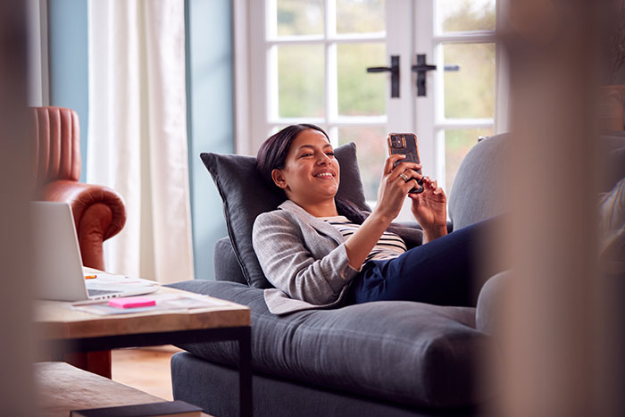 Woman relaxing on a couch smiling while looking at her smartphone, conveying a casual and comfortable moment.