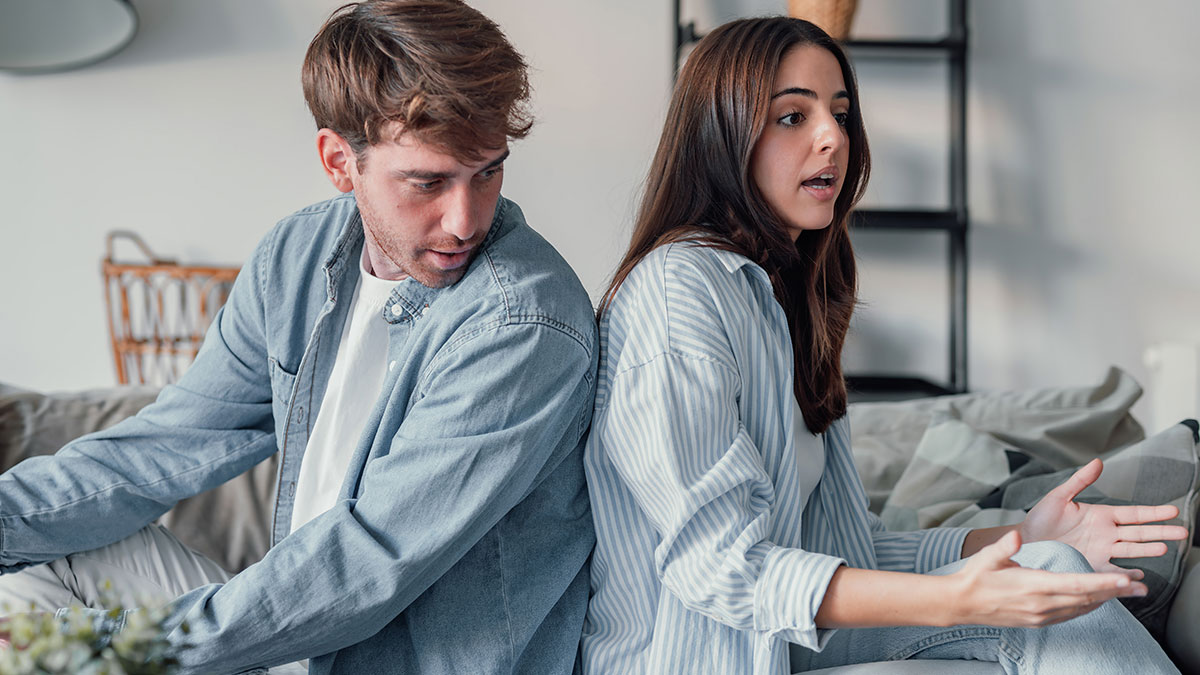 Young couple sitting back to back on couch, woman expressing frustration while man avoids eye contact, tense relationship moment.