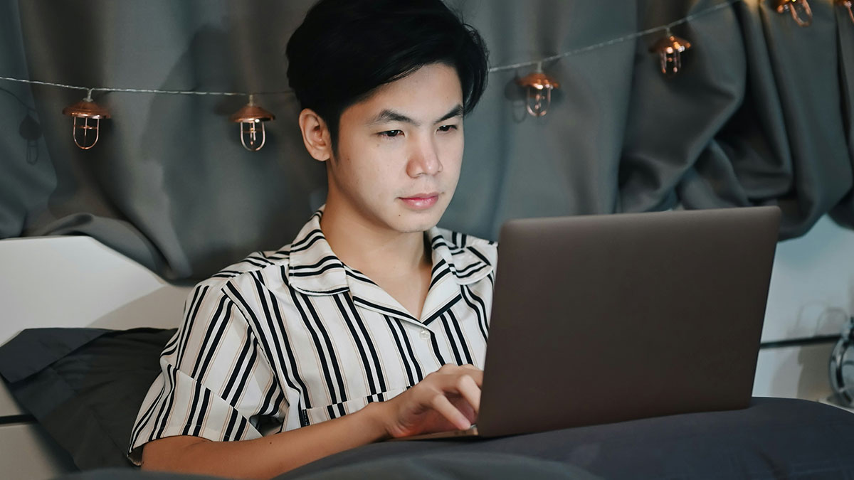 Young man in striped pajamas using laptop in bed, symbolizing creepy discovery of secret photos by girlfriend.
