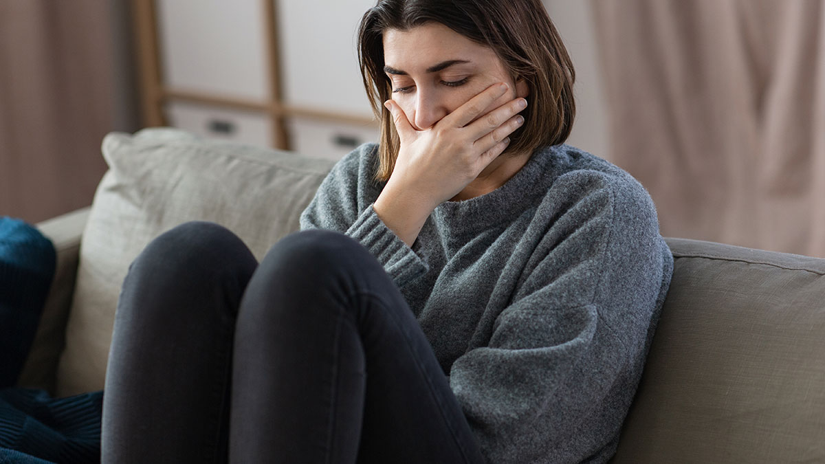 Young woman sitting on a couch, visibly upset and covering her mouth, after her boyfriend destroyed grandma's jewelry box.