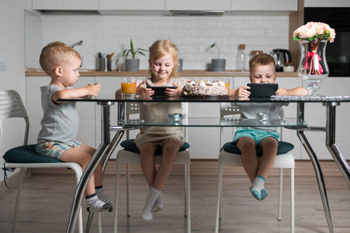 Three children sitting at a kitchen table, focused on tablets, with a cake and drinks nearby, reflecting family expectations. Three children sitting at a kitchen table, focused on tablets, with a cake and drinks nearby, reflecting family expectations.