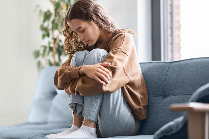 Young woman sitting curled up on a couch, looking upset and reflective in a quiet home setting.