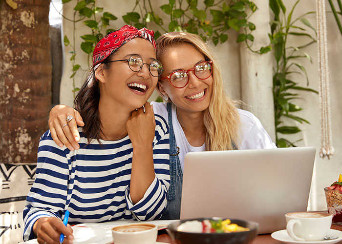 Two women smiling and looking at a laptop together, sharing a joyful moment in a cozy cafe setting. Two women smiling and looking at a laptop together, sharing a joyful moment in a cozy cafe setting.