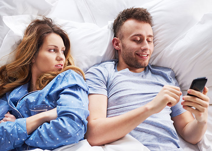 Couple in bed with woman upset and man smiling at phone, highlighting work wife sending hearts and mirror selfies. Couple in bed with woman upset and man smiling at phone, highlighting work wife sending hearts and mirror selfies.