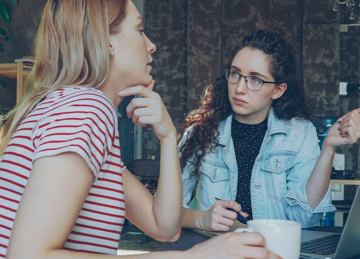 Two women having a serious conversation indoors, one wearing glasses and denim jacket, related to bridesmaid nails before wedding conflict