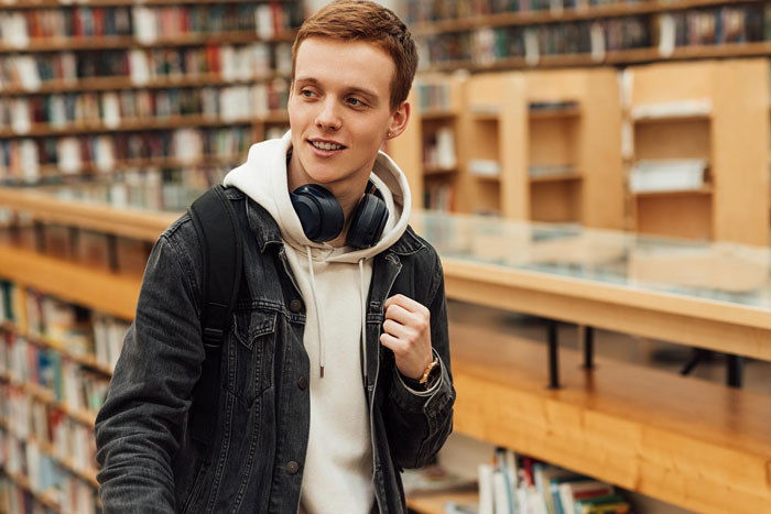 Young man in casual clothes with headphones and backpack in a library, representing a 23YO demanding allowance for doing nothing.