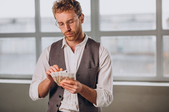 Young man wearing glasses and vest, counting money indoors, representing rich golden child demanding allowance while traumatized.