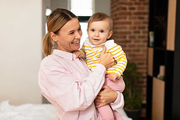 Woman holding baby smiling indoors, illustrating a live-in nanny job with challenging demands story context. Woman holding baby smiling indoors, illustrating a live-in nanny job with challenging demands story context.