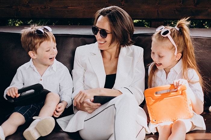 Woman in white suit wearing sunglasses, sitting with two kids holding tablets, reflecting rich folks and babysitter theme.