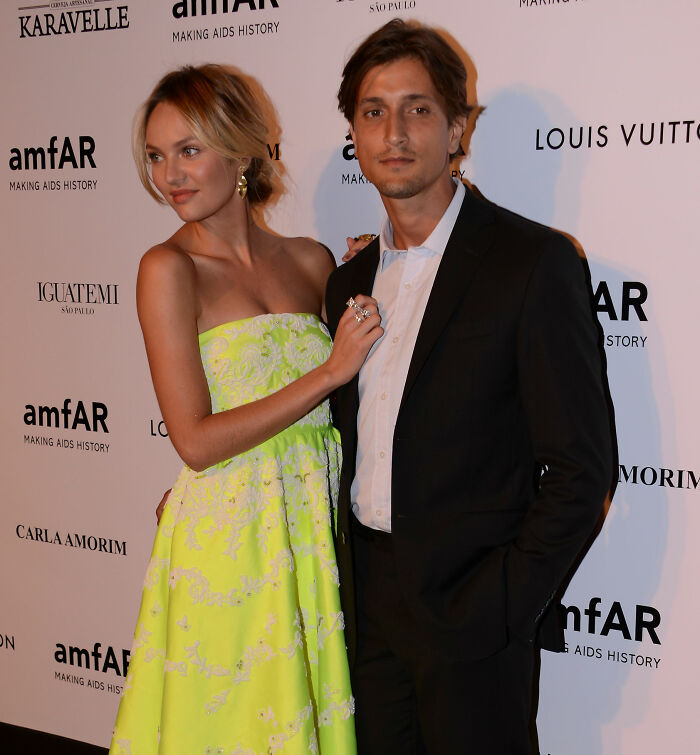 Victoria’s Secret model in a neon dress posing with a man in a black suit at a charity event backdrop. Victoria’s Secret model in a neon dress posing with a man in a black suit at a charity event backdrop.