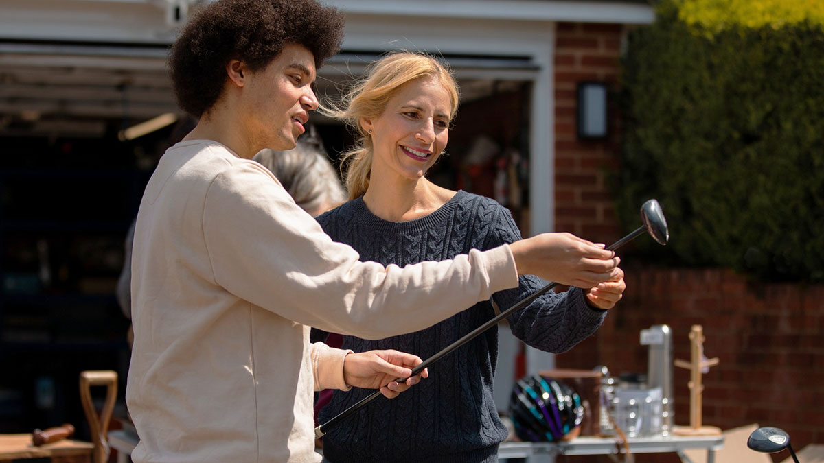A man and woman examining golf clubs outside a house, possibly negotiating a sale involving late hubbyu2019s clubs.