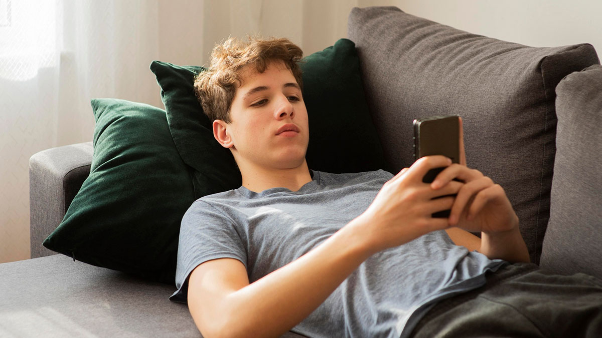 Young man lounging on sofa looking at phone, representing guy mooching off cousin and avoiding rent payment conflict.
