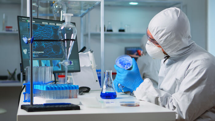 Scientist in protective suit examining a petri dish in a lab, highlighting people sharing potentially dangerous situations.