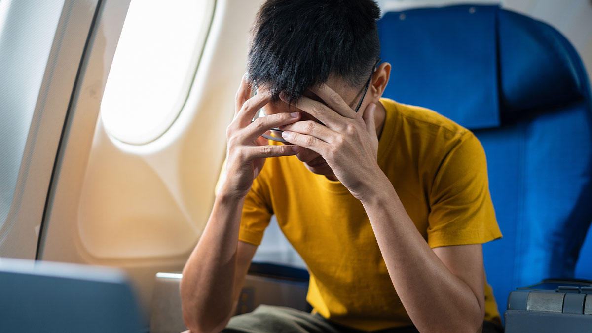 Man wearing glasses and yellow shirt stressed on a plane, covering face with hands near airplane window seat.