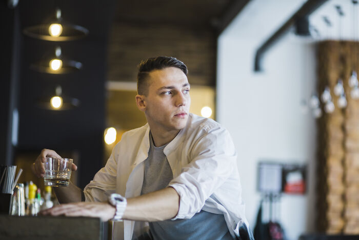 Young man sitting alone at a bar holding a glass, capturing the painful table for two moment turned table for one experience.