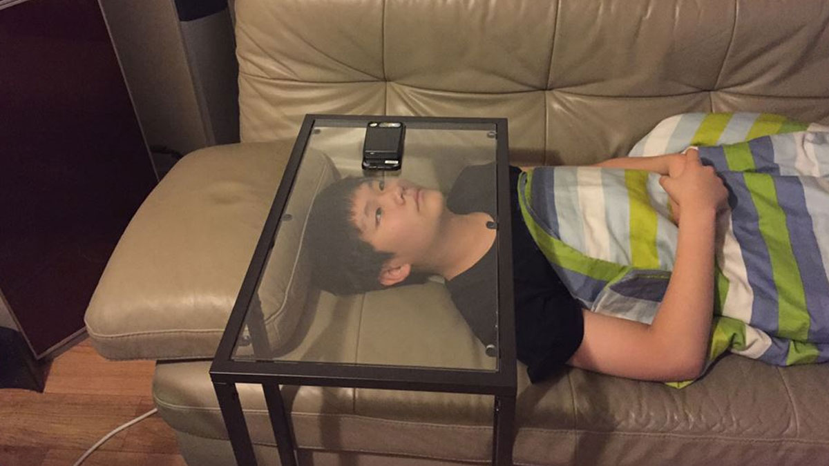 Boy using a creative invention with a glass table and phone holder while lying on a couch covered with a blanket.