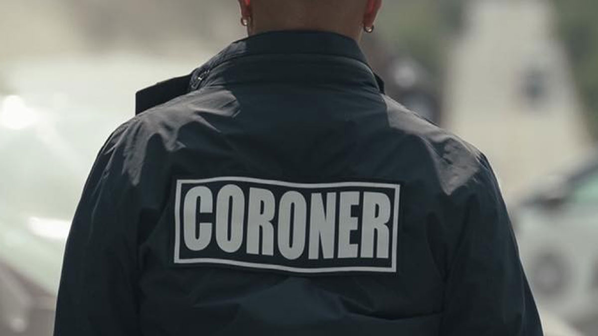 Person wearing a dark jacket with the word coroner on the back, standing outdoors in a blurred background.