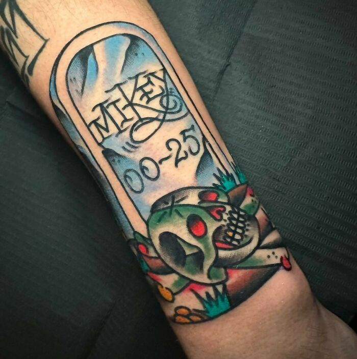 Tattoo of a gravestone with the name Miken and a green skull with red eyes on a person’s forearm.