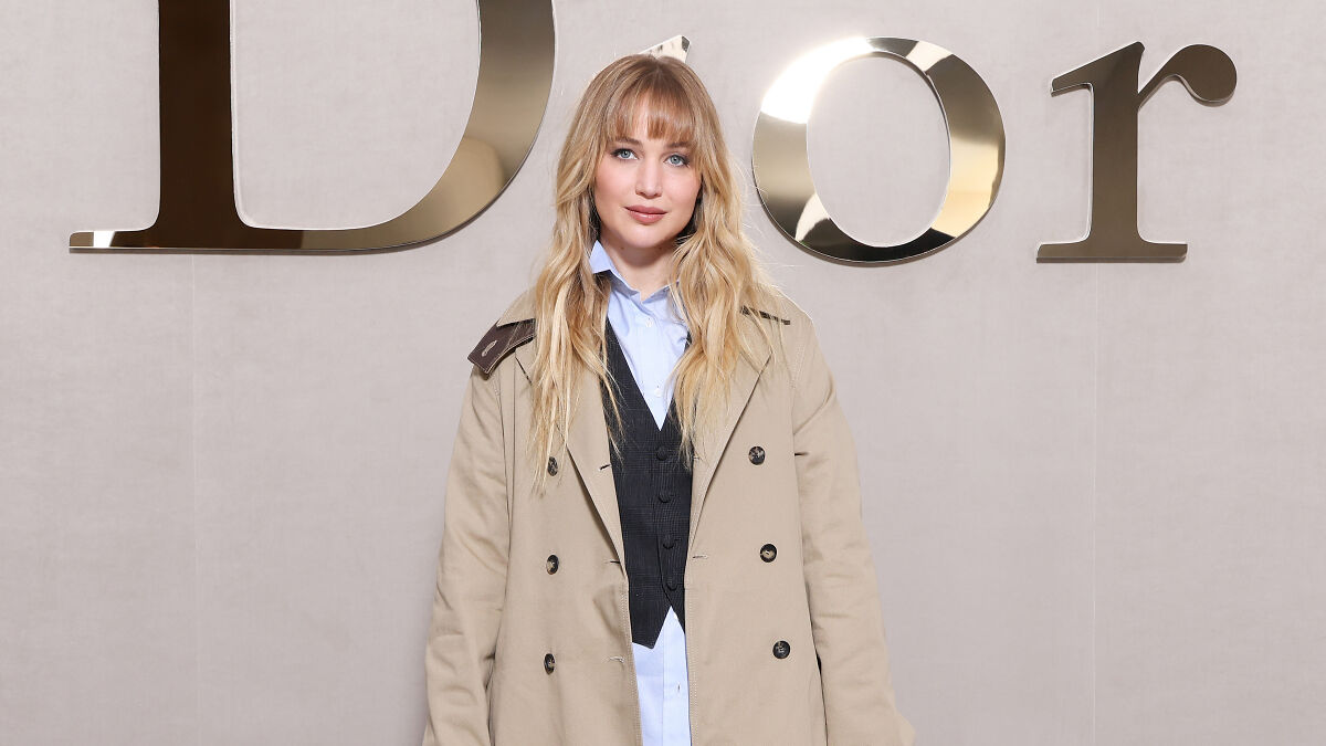 Blonde woman posing in beige coat and vest against a wall with large reflective Dior letters at Paris Fashion Week.