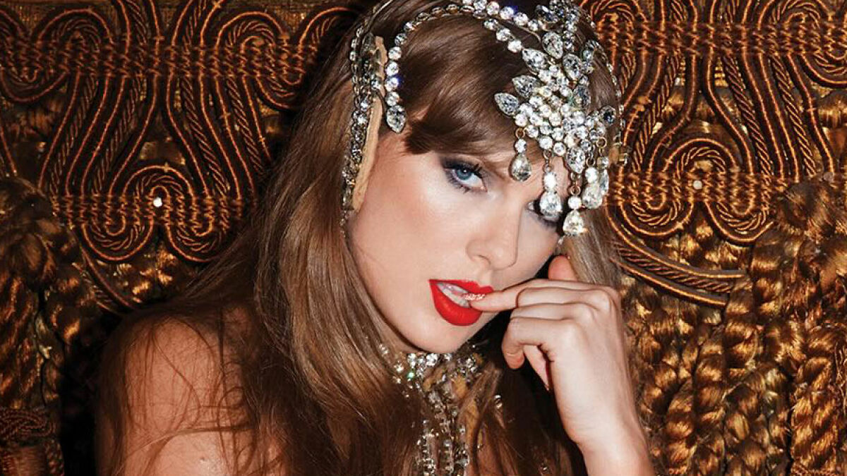 Taylor Swift wearing jeweled headpiece and red lipstick, posing with hand near mouth in ornate setting related to Travis Kelce manhood song.