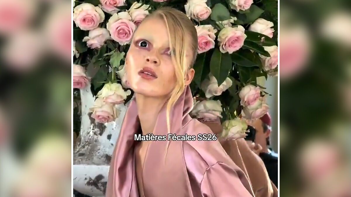Model with bold makeup and pink satin outfit posing in front of roses during Paris Fashion Week controversial show.