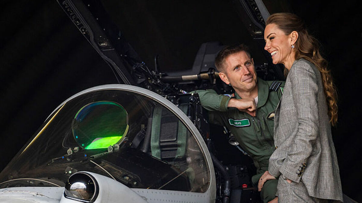 Servicemen seemingly checking out Kate Middleton as she smiles wearing a grey suit near an aircraft cockpit.
