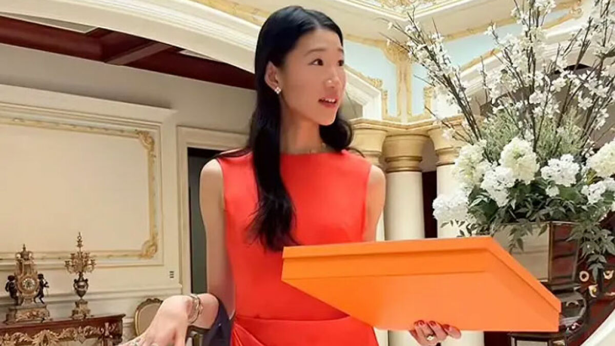 Billionaire influencer in a red dress holding an orange Hermu00e8s shopping box inside a luxurious room with floral decor.