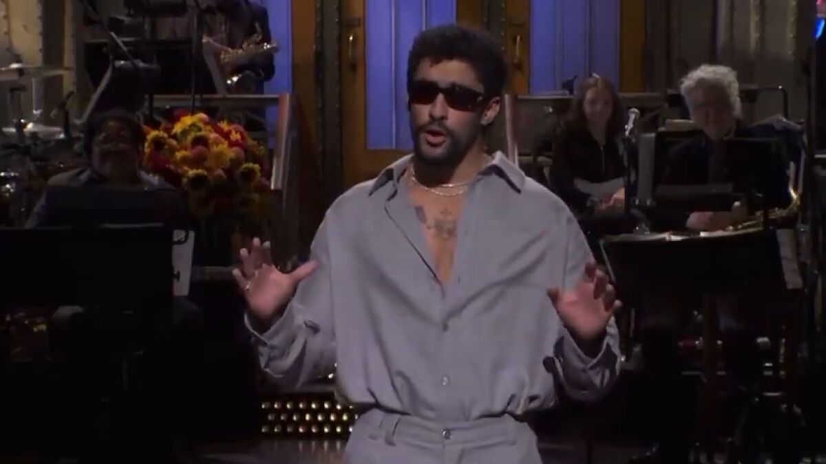 Bad Bunny delivering a monologue on SNL stage wearing sunglasses and a light gray outfit during a live performance.