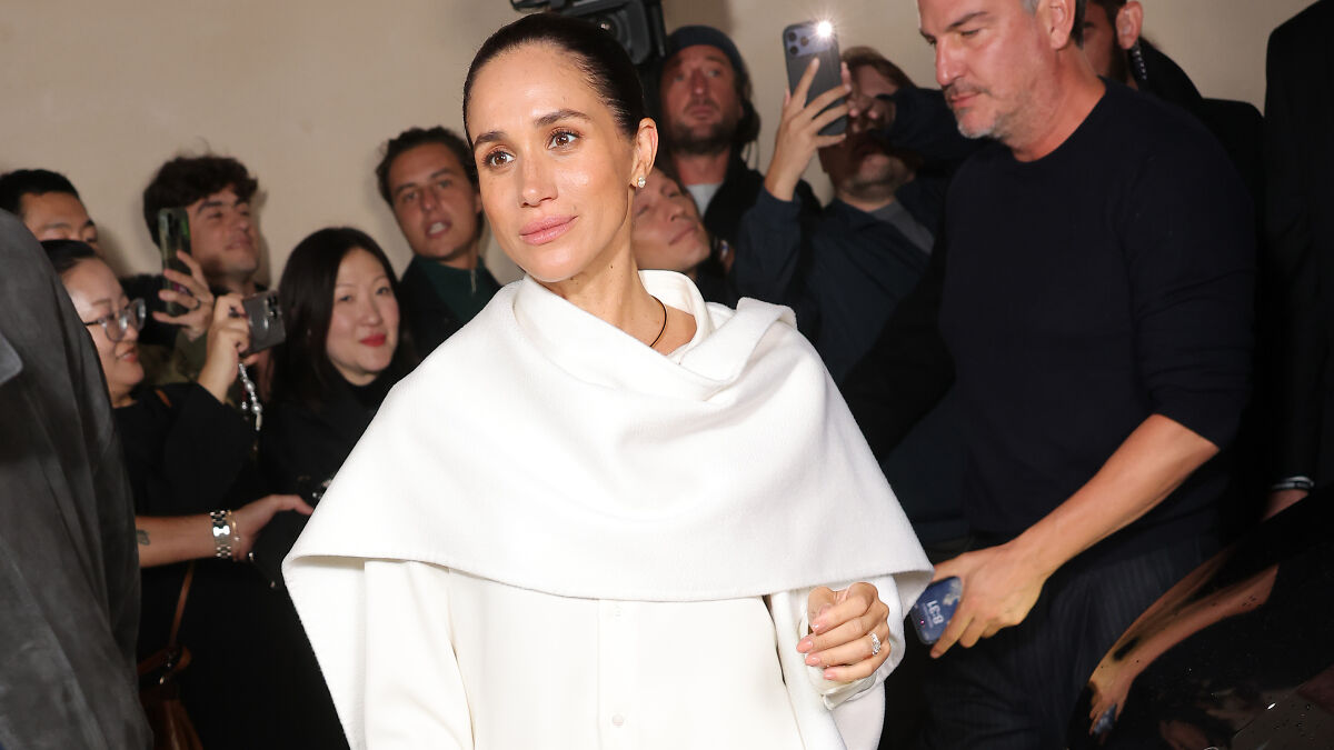 Meghan Markle in a white outfit arriving at Paris Fashion Week, surrounded by photographers and fans capturing her appearance.